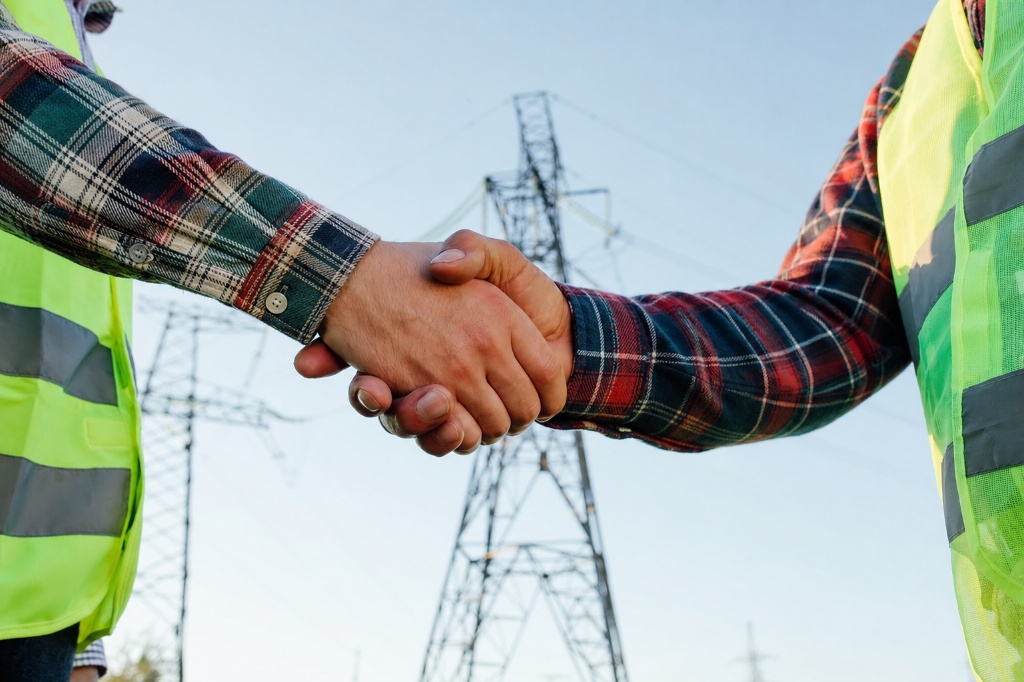 Two electrical workers in safety vests shaking hands in front of power transmission towers