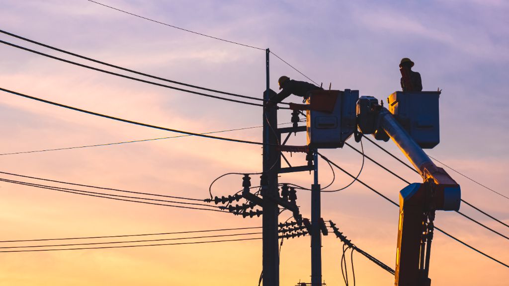 Utility linemen working from bucket trucks on power lines at sunset